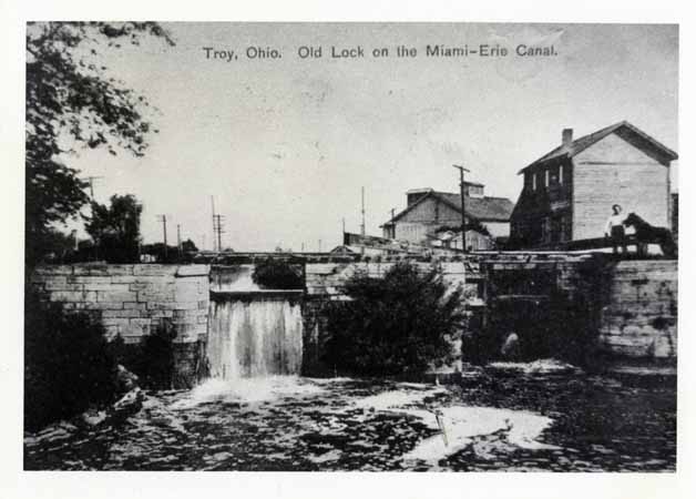A black and white postcard with a landscape image of a canal lock and a man standing to the right.