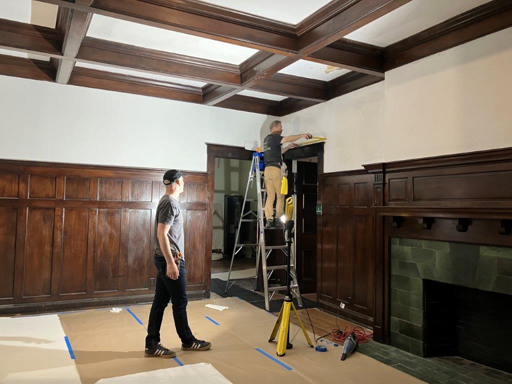 Two men one standing on a ladder working to install wallpaper liner in an empty room with wood paneling on the bottom portion of the wall.