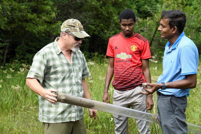 Three individuals standing in a meadow holding an auger and core sample they just removed.