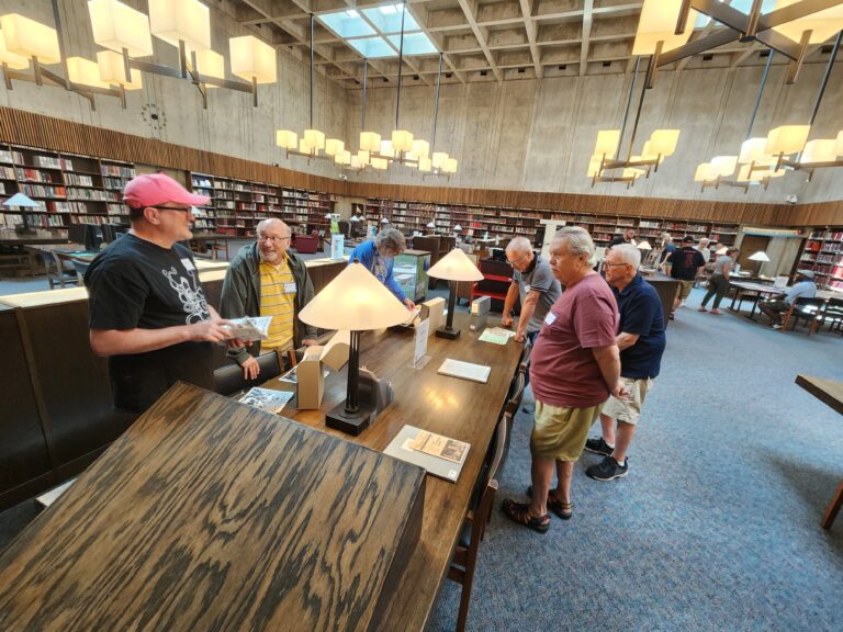 A photo of members of the Columbus Stonewall Union Trailblazers examining manuscripts from the GOHI Collection. The manuscripts are laid out on a wooden table in a large reading room with high ceilings and chandeliers.