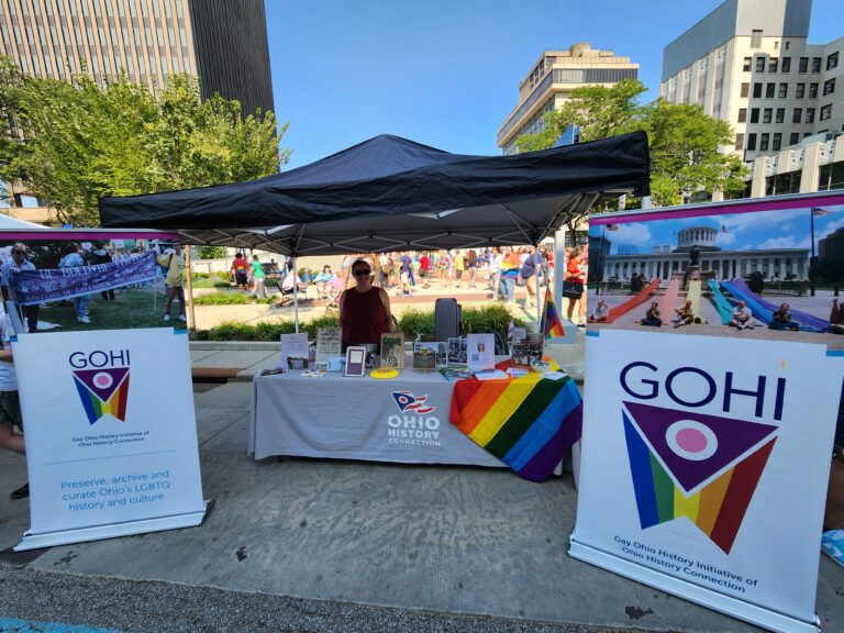 A photo of the GOHI table at Akron Pride 2024, including two banners with the GOHI logo and images from the archives. One of our GOHI staff is standing behind the table, smiling.