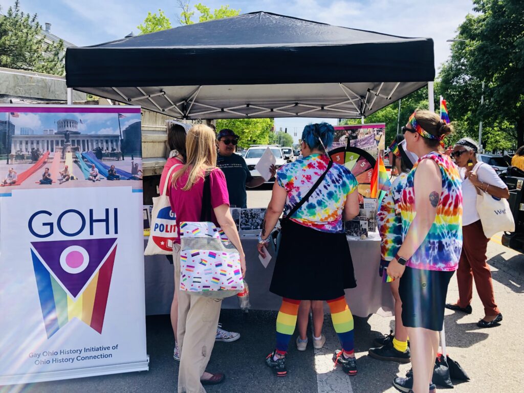 A photo of the GOHI table at Dayton Pride 2024, with several people in rainbow colors surrounding the table. There is a GOHI banner on the left side of the table.