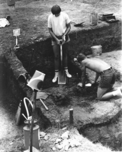 one student stands with a shovel while another student on their knees uses a trowel in a square excavation unit