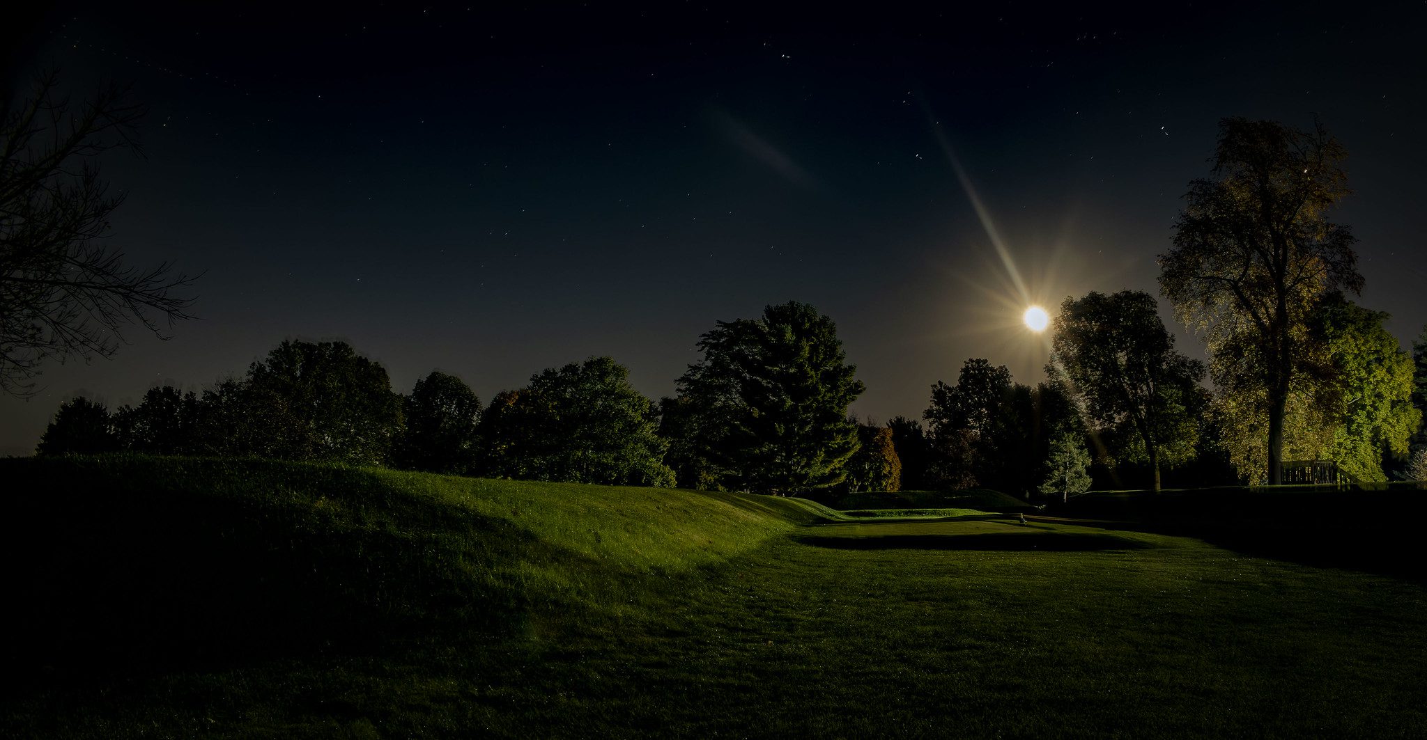 A flat space between two parallel earthen walls is in the foreground. The moon rises over a copse of trees in the background