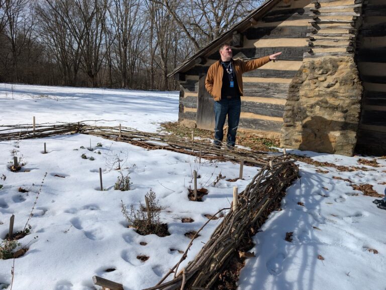 Man standing in from of a cabin wall pointing into the distance with a snow-covered garden area in the foreground