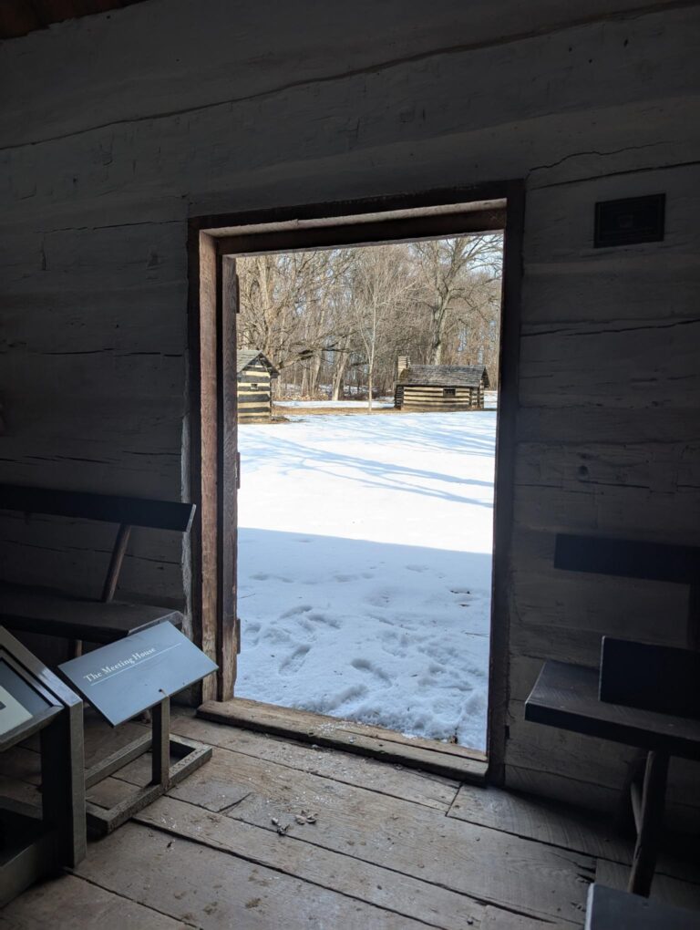 Picture taken from inside a cabin looking out an open doorway. The ground is covered with snow outside.